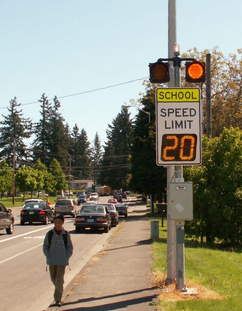 Student walking along sidewalk against traffic with large school speed limit sign and flashing lights